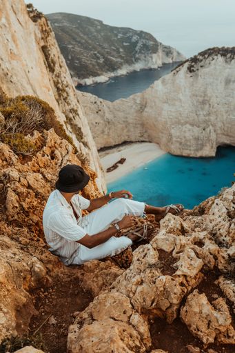 A back view of black man sitting on steep cliff overlooking the canyon in Greece