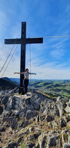 A vertical shot of a Caucasian woman standing in front of a cross located on Haselsteinwand mountain