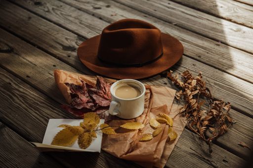 A high angle shot of a cup of coffee near a brown hat and dried leaves on a wooden surface