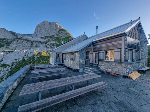 A mountain hut, with solar panels, and benches in front of it, located in Switzerland Alps, with a view on a rocky mountain in the background