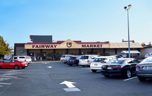 Fairway market grocery store and parking lot with numerous parked vehicles and white arrow directional signs painted on the asphalt.