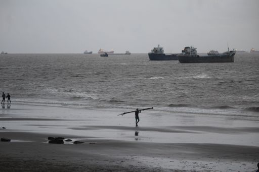 A grayscale shot of a fisherman holding fishing equipment walking on the beach with ships sailing in the background