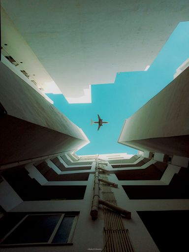 A low angle of a plane flying over white buildings seen in the bright blue sky