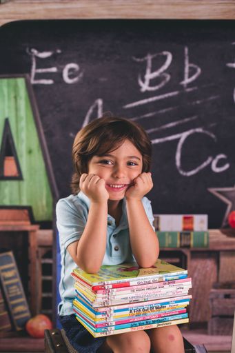 A vertical of a kid sitting with his elbows on books in front of the chalkboard.