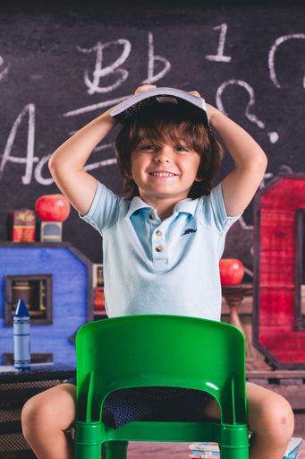 A little boy sitting on a green chair with a blue t-shirt is holding a notebook to his head in the classroom