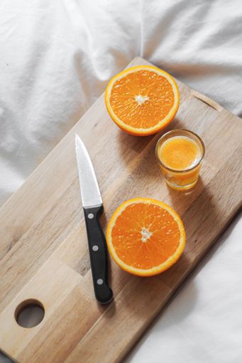 A vertical shot of a glass of juice and a sliced orange with a knife on a wooden board on white sheets