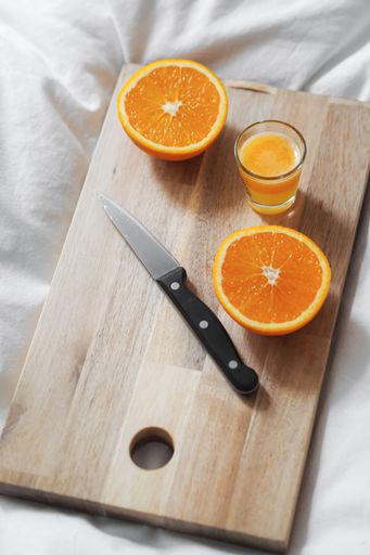 A vertical shot of a glass of juice and a sliced orange with a knife on a wooden board on white sheets