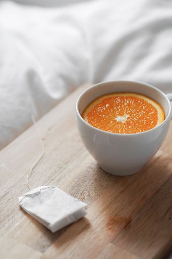 A vertical shot of a tea cup with a sliced orange in it next to a tea bag on a wooden board on white fabric