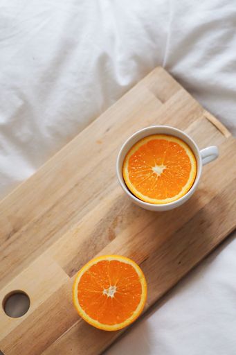 A vertical shot of a teacup with orange slice in it next to an orange cut on a wooden board on white sheets