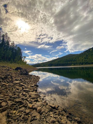 A vertical shot of a lake surrounded by green pine trees under a cloudy blue sky