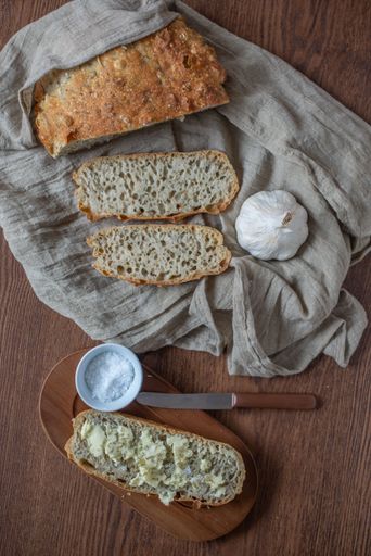 A vertical top view of pieces of no-knead bread, a head of garlic and salt on a wooden tray