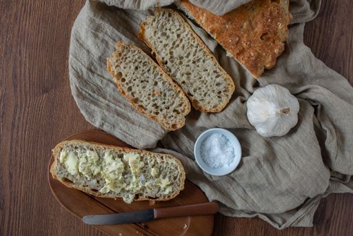 A top view of pieces of no-knead bread on a towel, a head of garlic and a knife on a tray