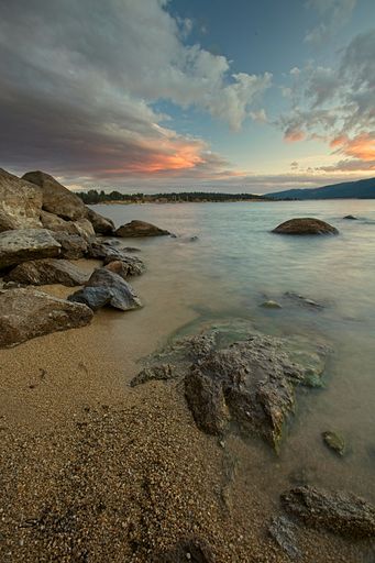 A vertical shot of the sandy beach with rocks and the calm lake with the sunset in the background