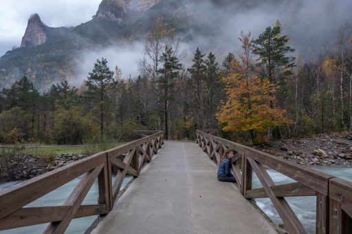 A woman sitting on a bridge with wooden railings against the background of autumn trees and misty mountains