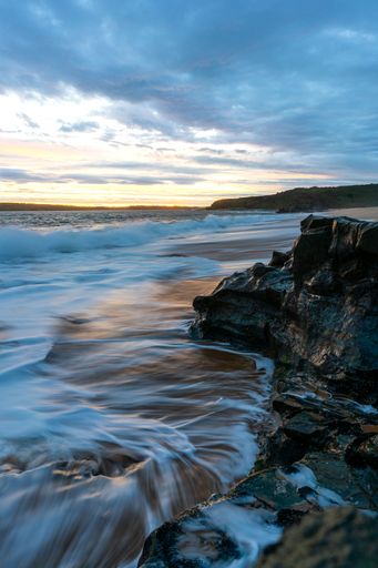 A vertical shot of a sea with waves approaching the cliffs of the beach and the sunset on the horizon