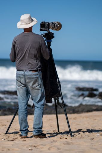 A surf photographer taking photos with a telephoto zoom lens and camera body on a tripod