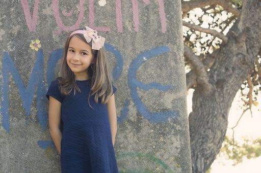 A closeup of a cute little girl in a blue dress leaning against a wall.