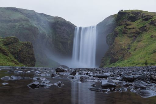 A low-angle of Skogafoss waterfall flowing with a long exposure and making a pond, hills covered with meadow and cloudy sky background