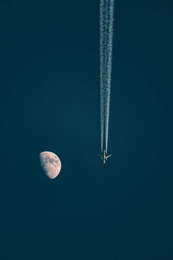 A vertical shot of a plane flying against a dark blue sky with a half Moon in the background