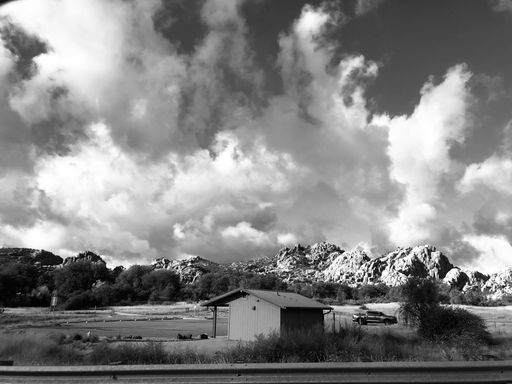 A breathtaking grayscale shot of fluffy cloudscape over rural field with small building and rocky hills