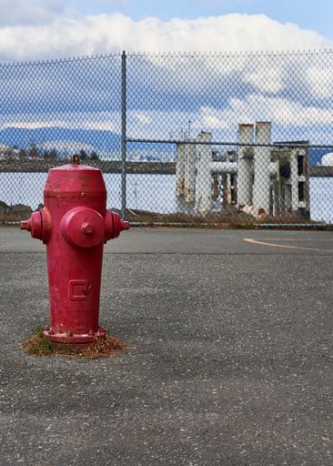 A close up of a red fire hydrant in a parking lot beside the ocean and ferry docking pilings and white fluffy clouds in the background.