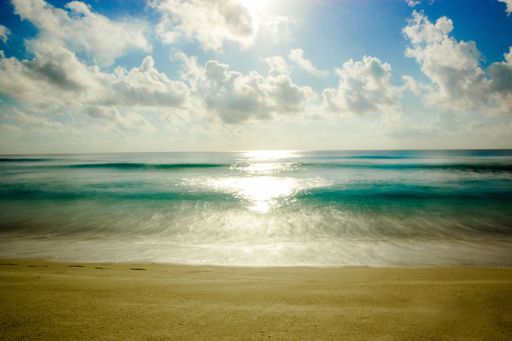 A blue ocean with a sandy beach and sunlight reflected in the waters under blue sky