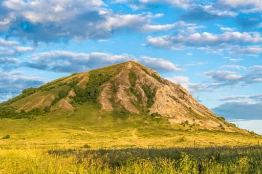 Summer landscape with green pasture, ancient relic mountain Shihan, blue cloudy sky in rays of setting sun. Golden hour.
