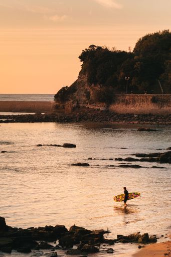 A vertical shot of a man surfing in the sea in Spain under the colorful sunset, the beach with trees and rocks