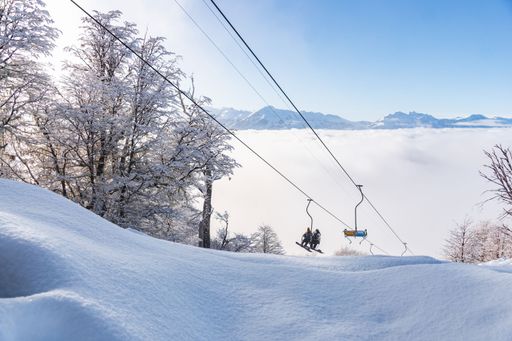 A scenic view of a ski lift surrounded by a snow bank, trees, and mountains in the background