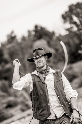 A vertical grayscale shot of a male wearing a cowboy outfit while whipping