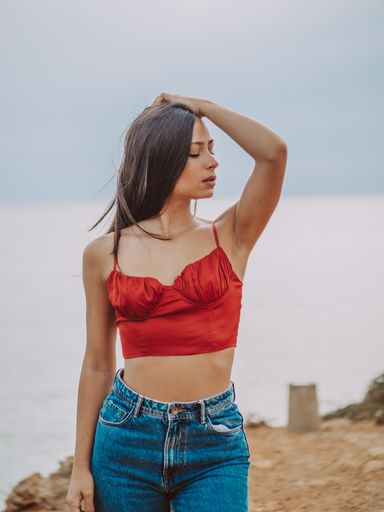 A beautiful young female wearing a sexy red crop top and jeans and standing on a cliff with the sea in the background