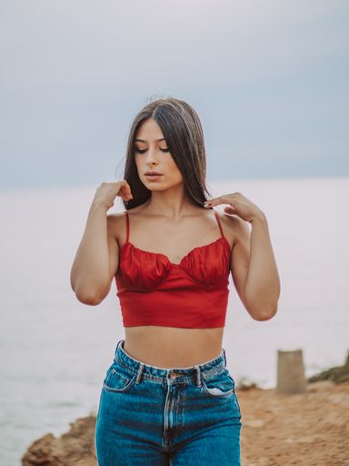 A beautiful young female wearing a sexy red crop top and jeans and standing on a cliff with the sea in the background