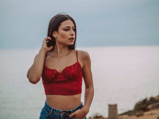 A beautiful young female wearing a sexy red crop top and jeans and standing on a cliff with the sea in the background