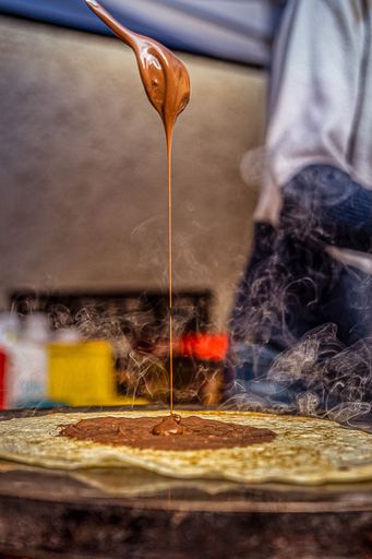 A vertical closeup of a spoon with chocolate pouring down to the hot blini