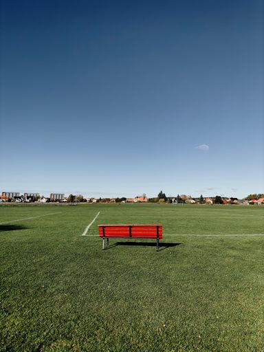 A vertical shot of an empty red bench on the green grass under a blue sky