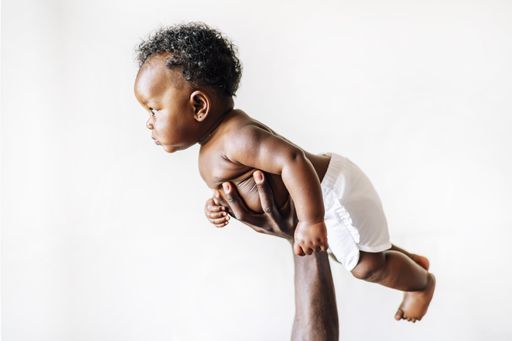 An African-American baby being held up in the air by her parent against a white wall