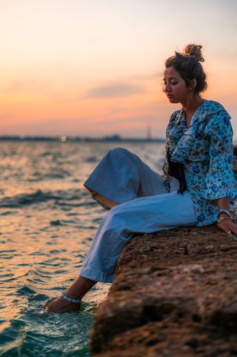 A young female wearing her hair in a messy bun and relaxing on stones surrounded by the sea during the sunset