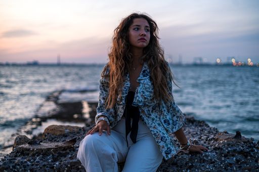 A shallow focus of a stylish female with messy curly hair sitting on rocks surrounded by the sea in the evening