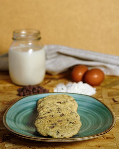 A vertical shot of chocolate chip cookies next to a jar of milk, with eggs and flour on a wooden surface