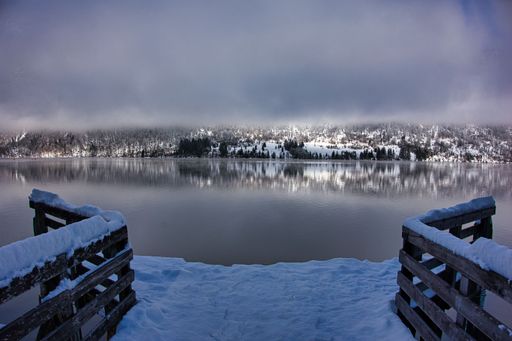 A scenic shot of a calm lake with trees behind it seen from a snow-covered wooden dock