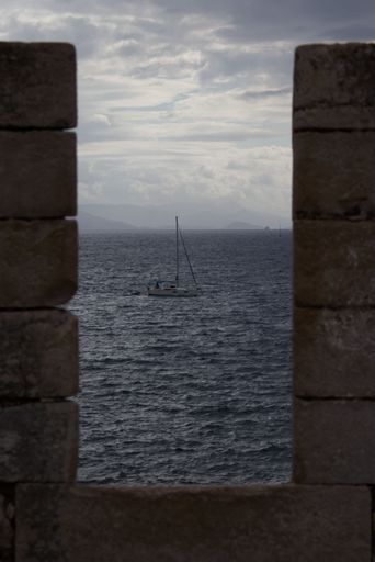 A vertical shot of a sailing boat framed between the walls of the old Corfu fort on a cloudy day