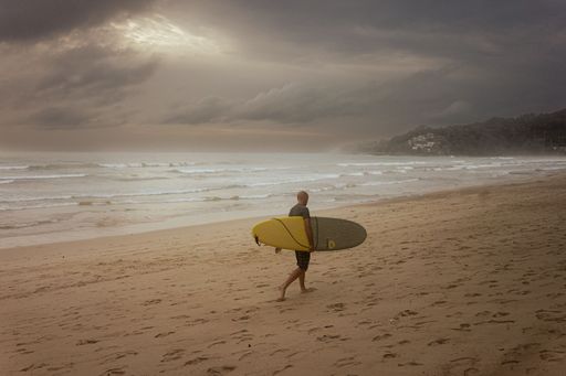 The man with a surfboard walking along the sandy beach on a cloudy day.