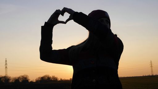 A silhouette of a female shaping a heart with her hands against the sky at sunset