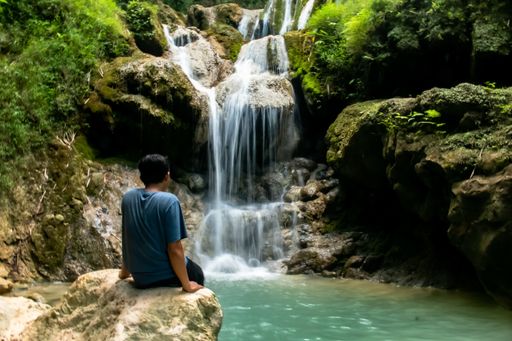 Backside view from a young Asian man sitting on a rock and looks on the waterfall Mudal river, Java, Indonesia