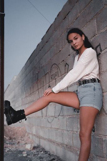 A vertical shot of a cool caucasian female in jeans shorts and boots leaning on a stone wall