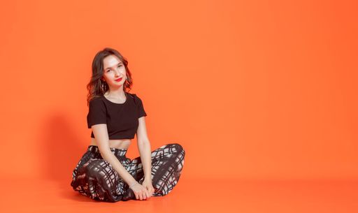 Young elegant brunette girl in black pantsuit sits on orange background. She lowered her hands in front of her. Looks at camera. Studio photography.