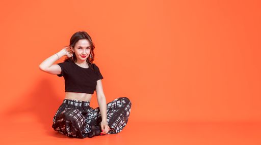 Young elegant brunette girl in black pantsuit sits on orange background. She put one hand behind her head. Looks at camera. Studio photography.
