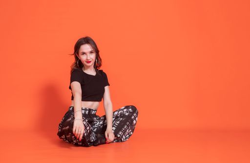 Young elegant brunette girl in black pantsuit sits on orange background. She folded her hands on knees. Looks at camera. Studio photography.