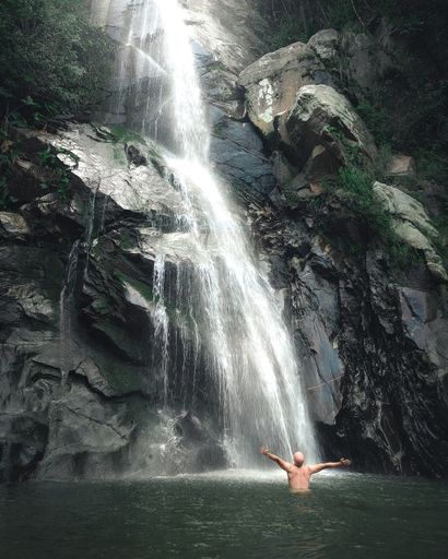 A vertical shot of a naked man in the lake against a scenic waterfall