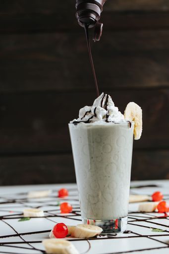 A closeup shot of chocolate drizzling over a tall glass cup of milkshake topped with whipped cream